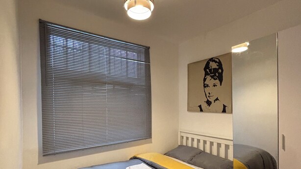 Ein Schlafzimmer mit einem Bett, einem Fenster mit Jalousien, einer Deckenleuchte, einem an der Wand befestigten Spiegel und einem Holzboden.