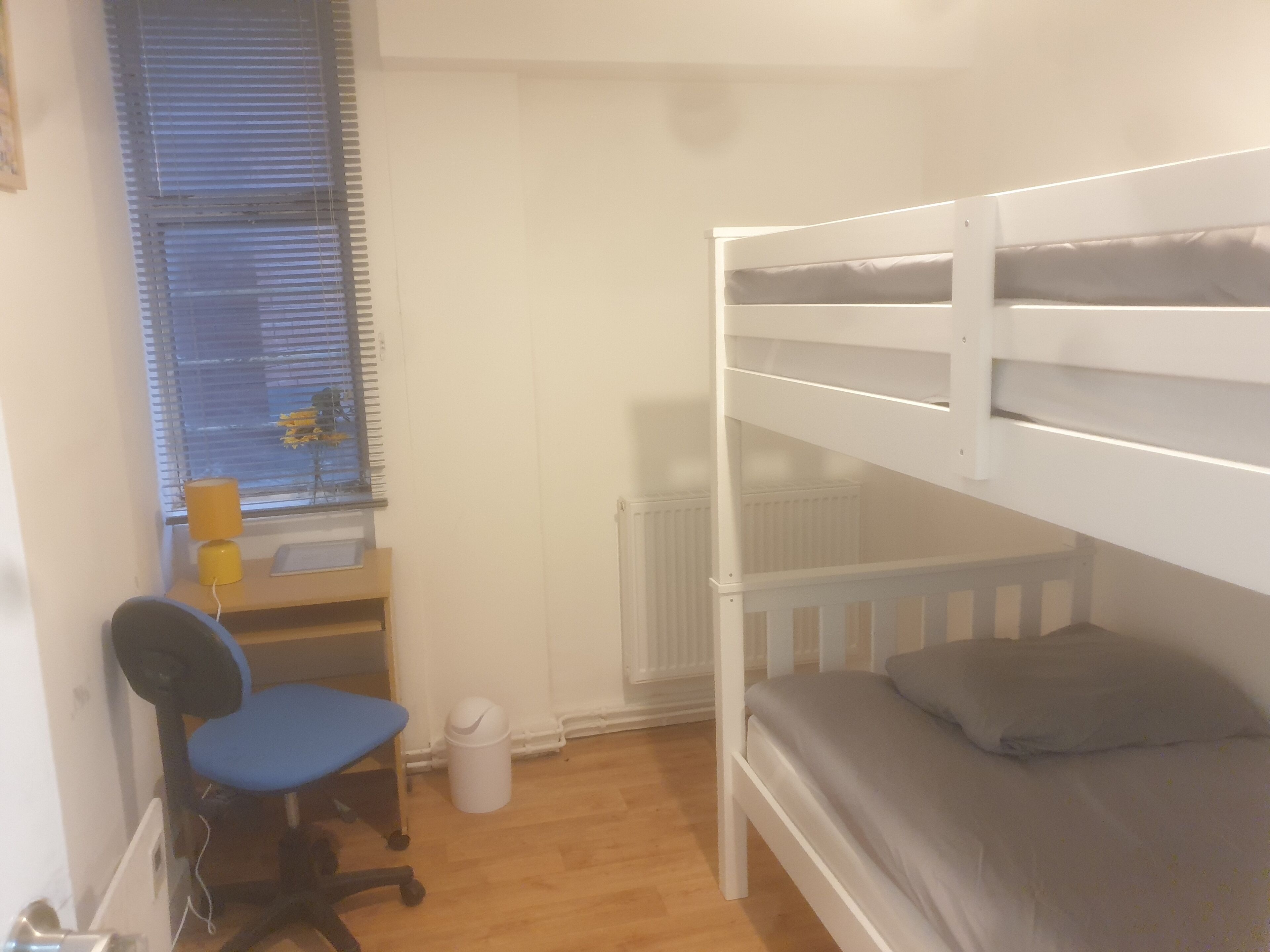 A bunk bed room with a desk and chair, a radiator, and a window with blinds.