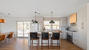 Une cuisine moderne avec des meubles blancs, un îlot central doté de tabourets, un coin repas avec une table et des chaises en bois, et des luminaires suspendus.