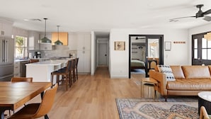 Une cuisine moderne avec des meubles blancs, un îlot central et des luminaires suspendus. On y trouve un coin repas avec une table et des chaises en bois, ainsi qu’un salon avec un canapé en cuir et un tapis à motifs.