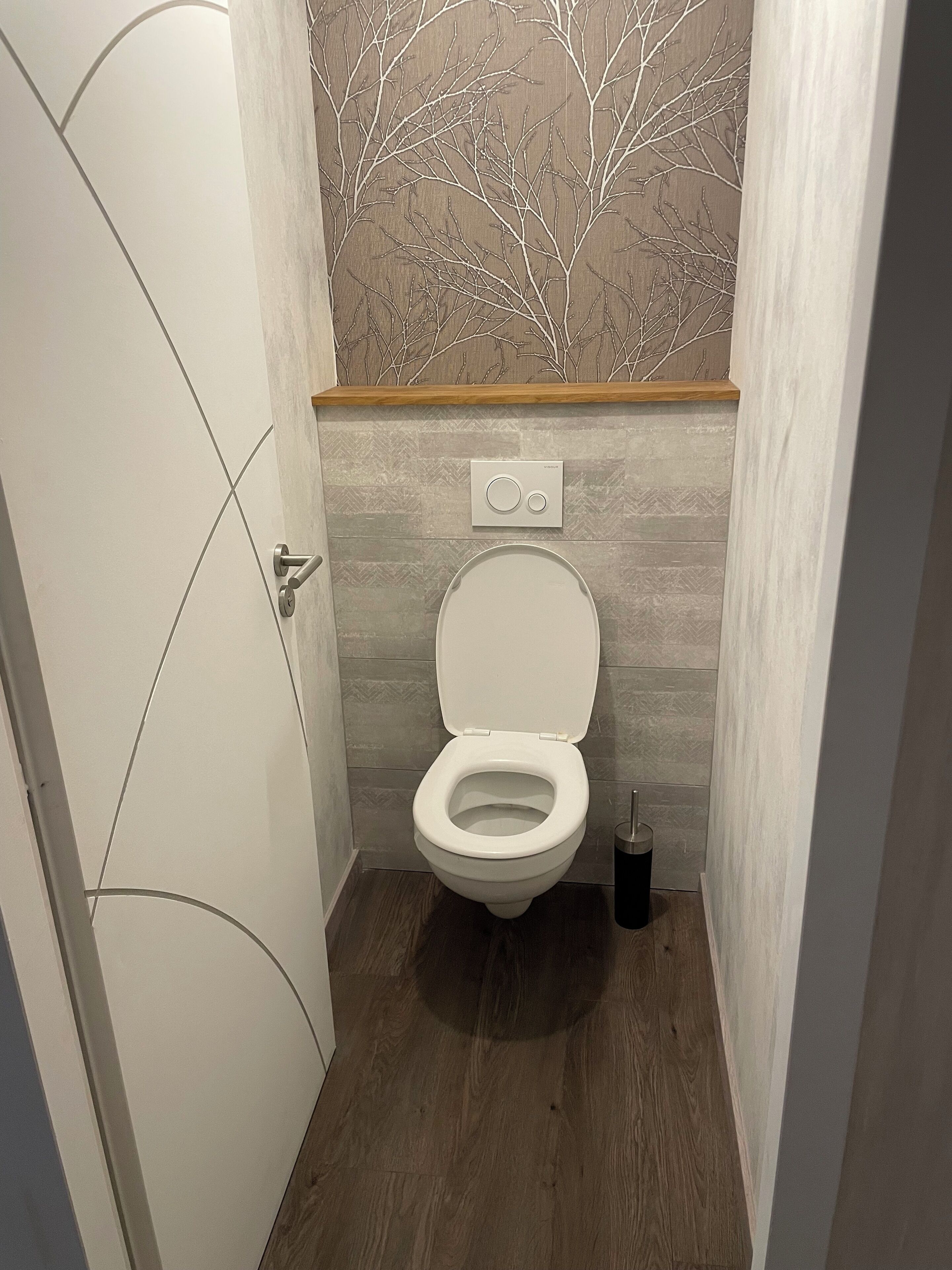 Een kleine badkamer met toilet, een bediend hendel aan de muur en een houten plank boven het toilet.