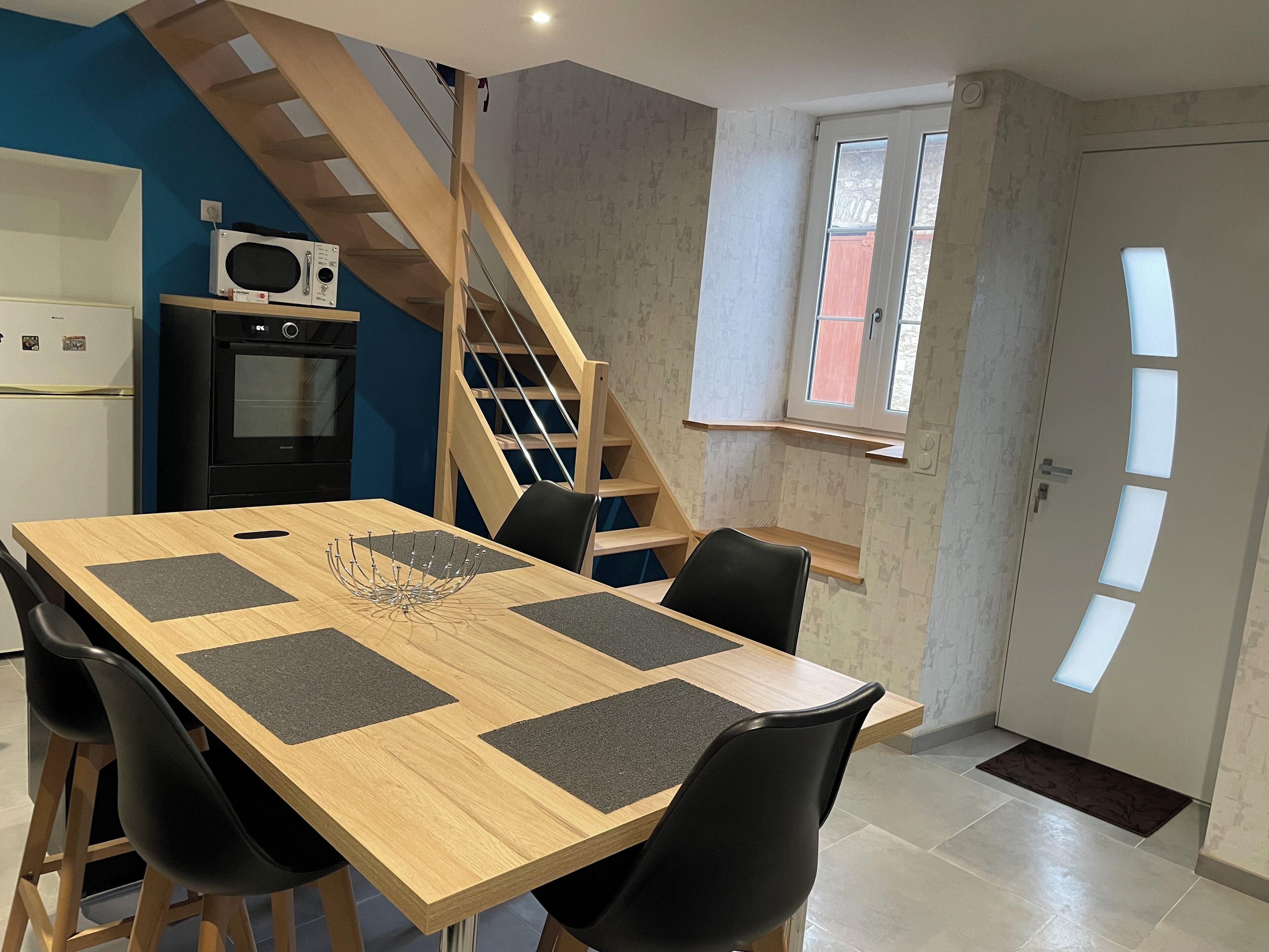 A dining area with a wooden table, black chairs, a decorative centerpiece, a kitchen with a microwave and oven, a wooden staircase, and a window with a view.