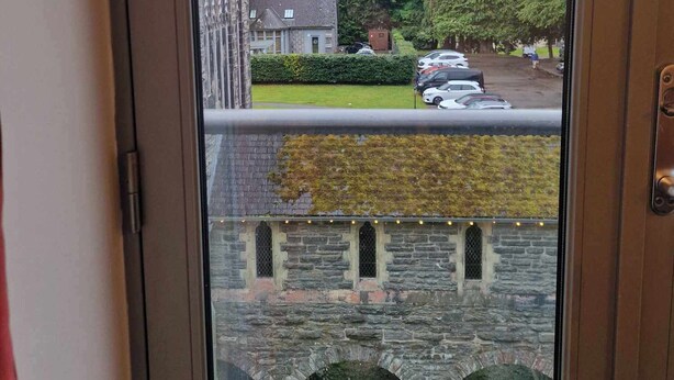 Vista a través de una ventana de un edificio de piedra, con un área verde y coches aparcados al frente.