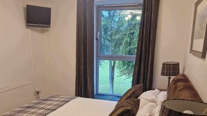 Un dormitorio con una cama grande, una mesita de noche de madera, una lámpara, un televisor y una ventana con cortinas.