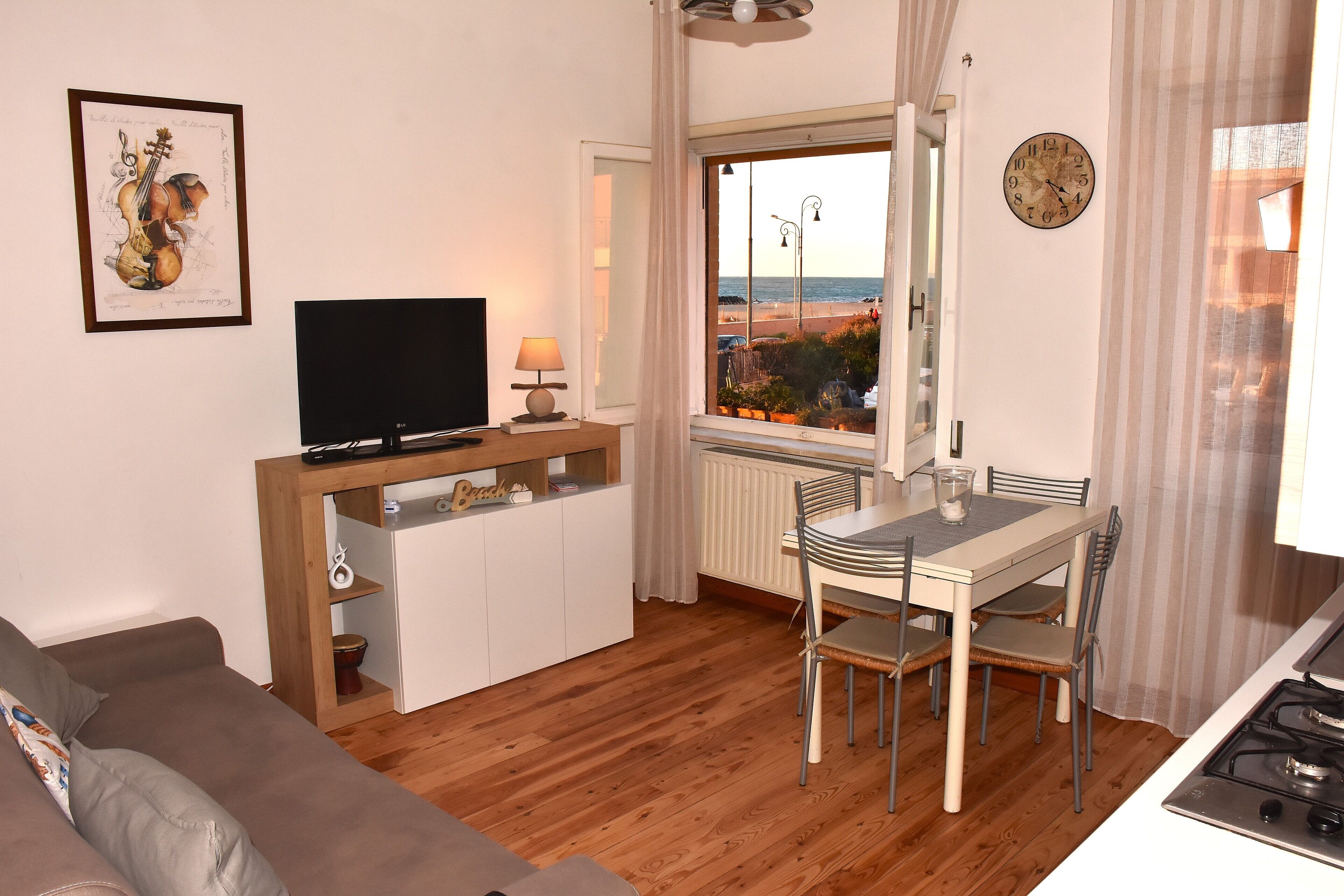 A living room with a TV, a dining table with chairs, a sofa, a window with a view of the beach, and a clock on the wall.