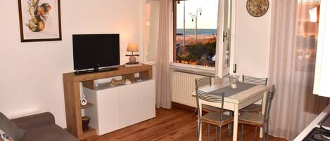 A living room with a TV, a dining table with chairs, a sofa, a window with a view of the beach, and a clock on the wall.