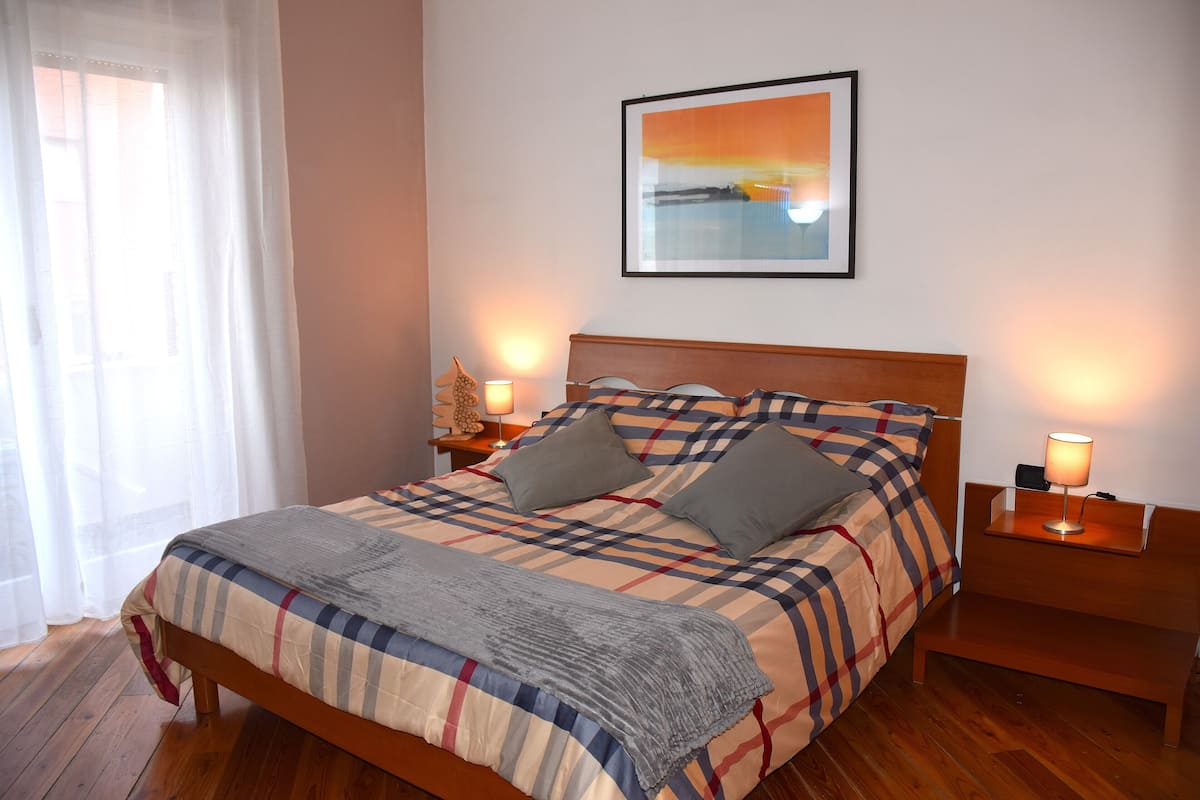 Ein ordentlich bezogenes Bett mit kariertem Überwurf und Kissen, ein Nachttisch mit Lampe und ein gerahmtes Bild an der Wand.