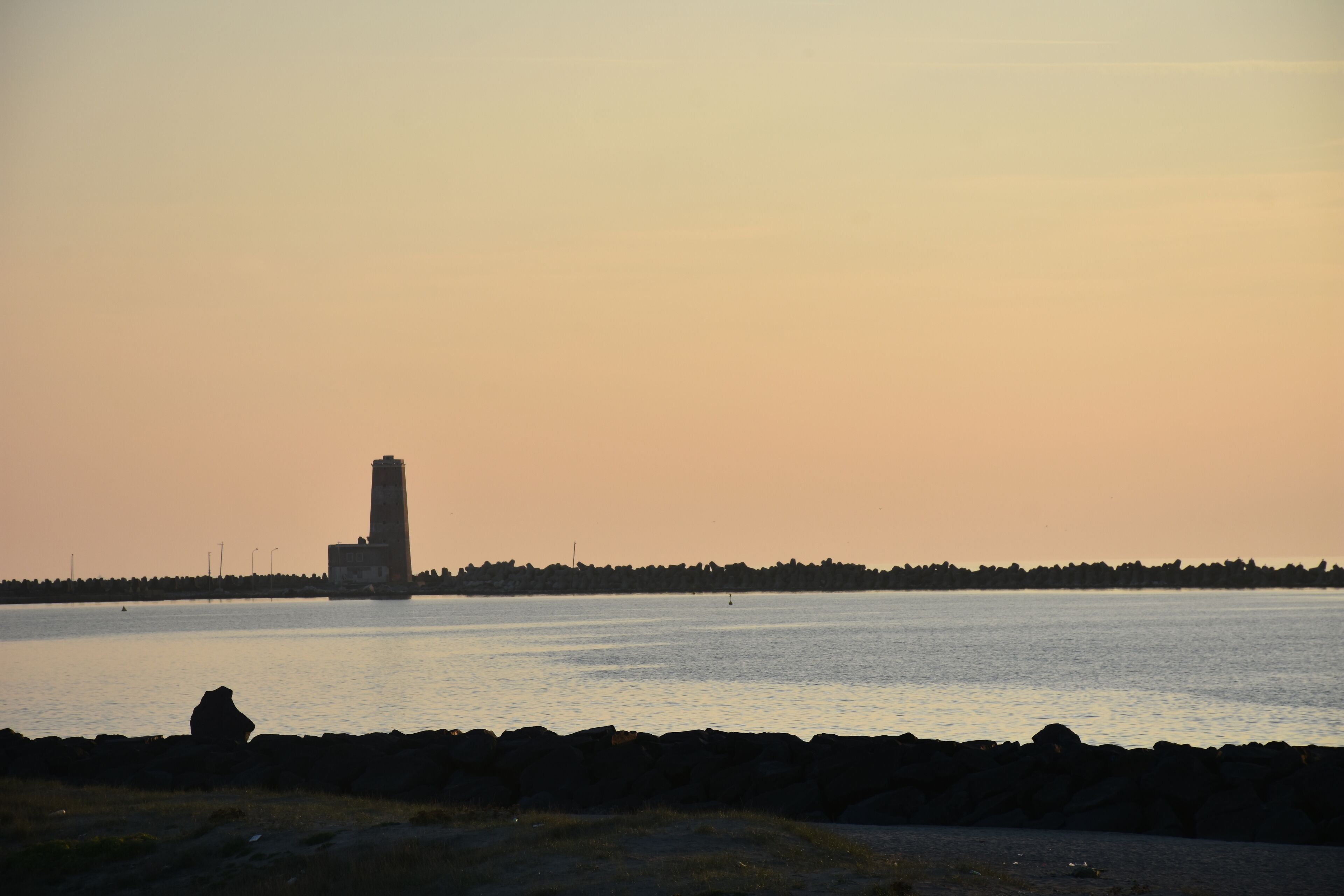 Escena costera con un faro, costa rocosa y una persona sentada sobre las rocas.