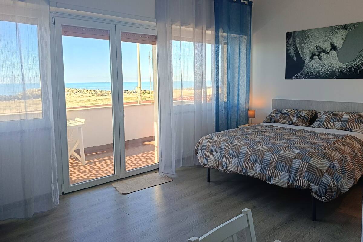 Ein Schlafzimmer mit Bett, Nachttisch, einem Gemälde und Blick auf den Strand durch eine Schiebetür.