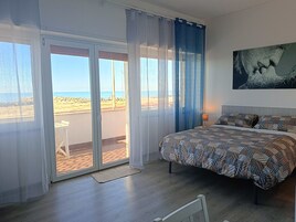 Et soveværelse med seng, et natbord, et maleri og udsigt til stranden gennem en skydedør.