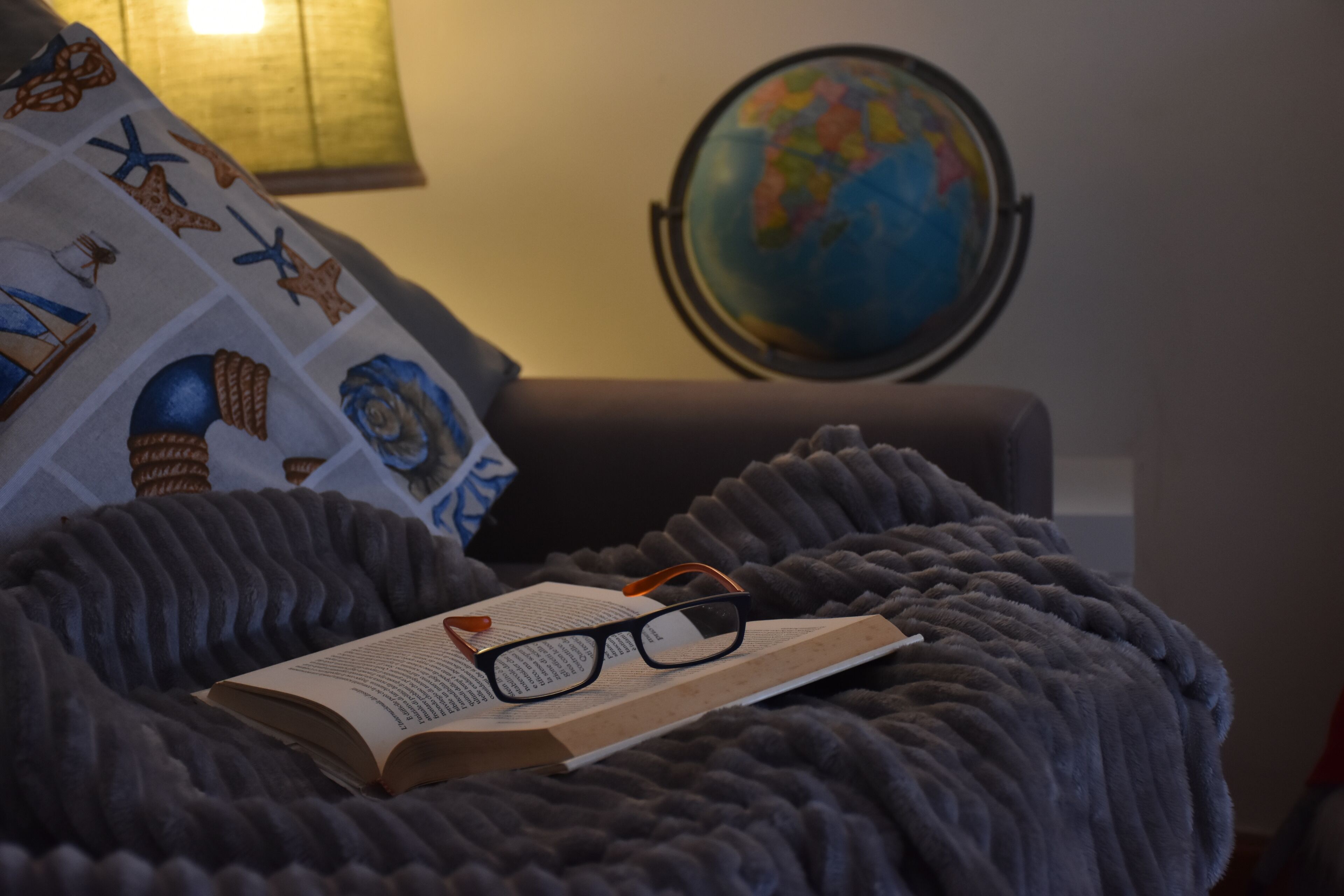 A cozy reading nook with a plush blanket, a book, and glasses on a chair. A globe and a pillow with nautical designs are in the background.