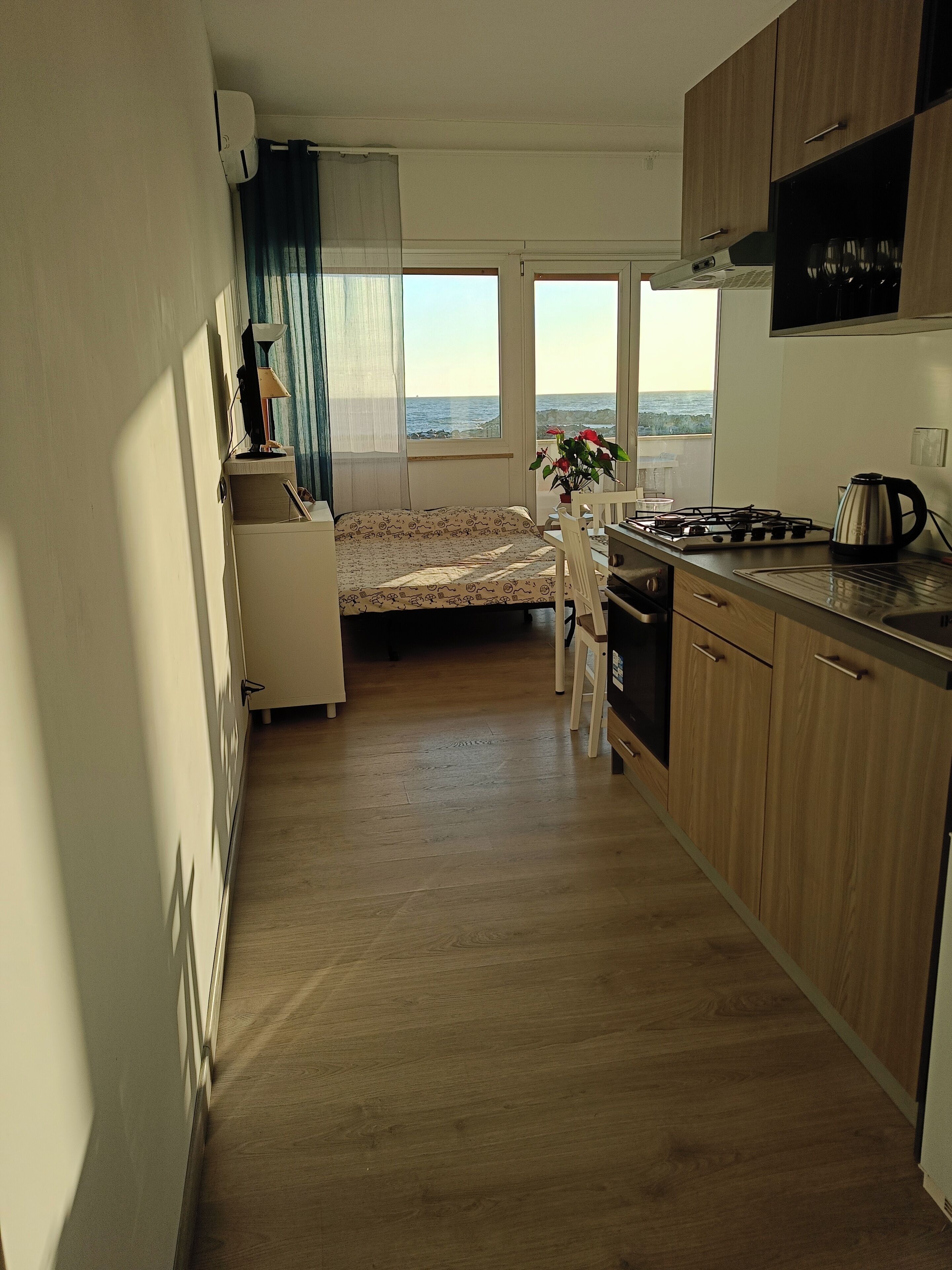Cocina compacta con una cama al fondo, vista al mar y zona de comedor con mesa y sillas.