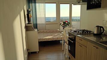Cocina compacta con una cama al fondo, vista al mar y zona de comedor con mesa y sillas.