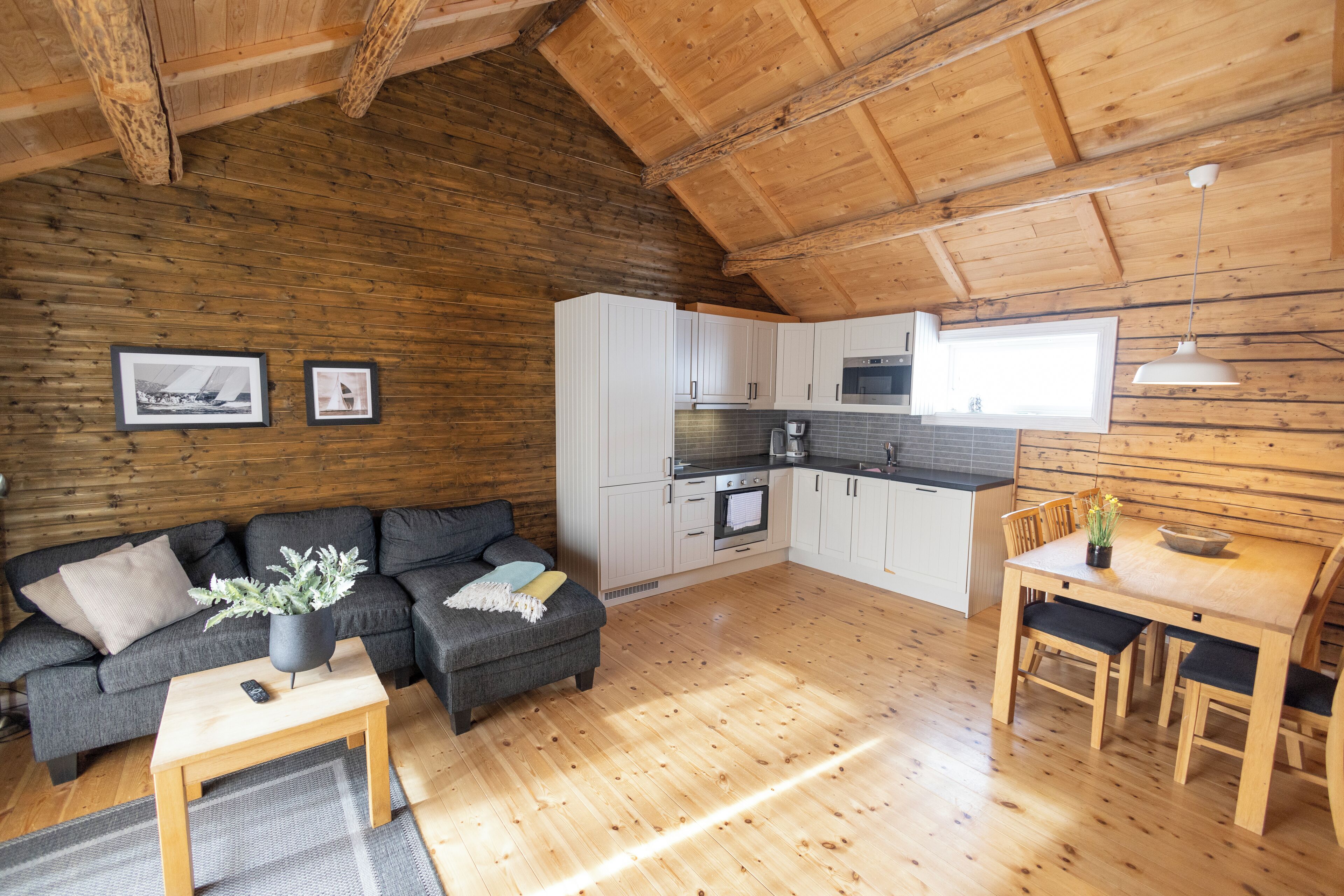 Un salon chaleureux, doté d’un plancher en bois, avec un canapé, une petite table et une table à manger avec des chaises. La cuisine est équipée de meubles blancs et d’un four à micro-ondes.