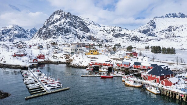 Un pueblo costero con edificios nevados, un muelle con barcos y montañas al fondo.