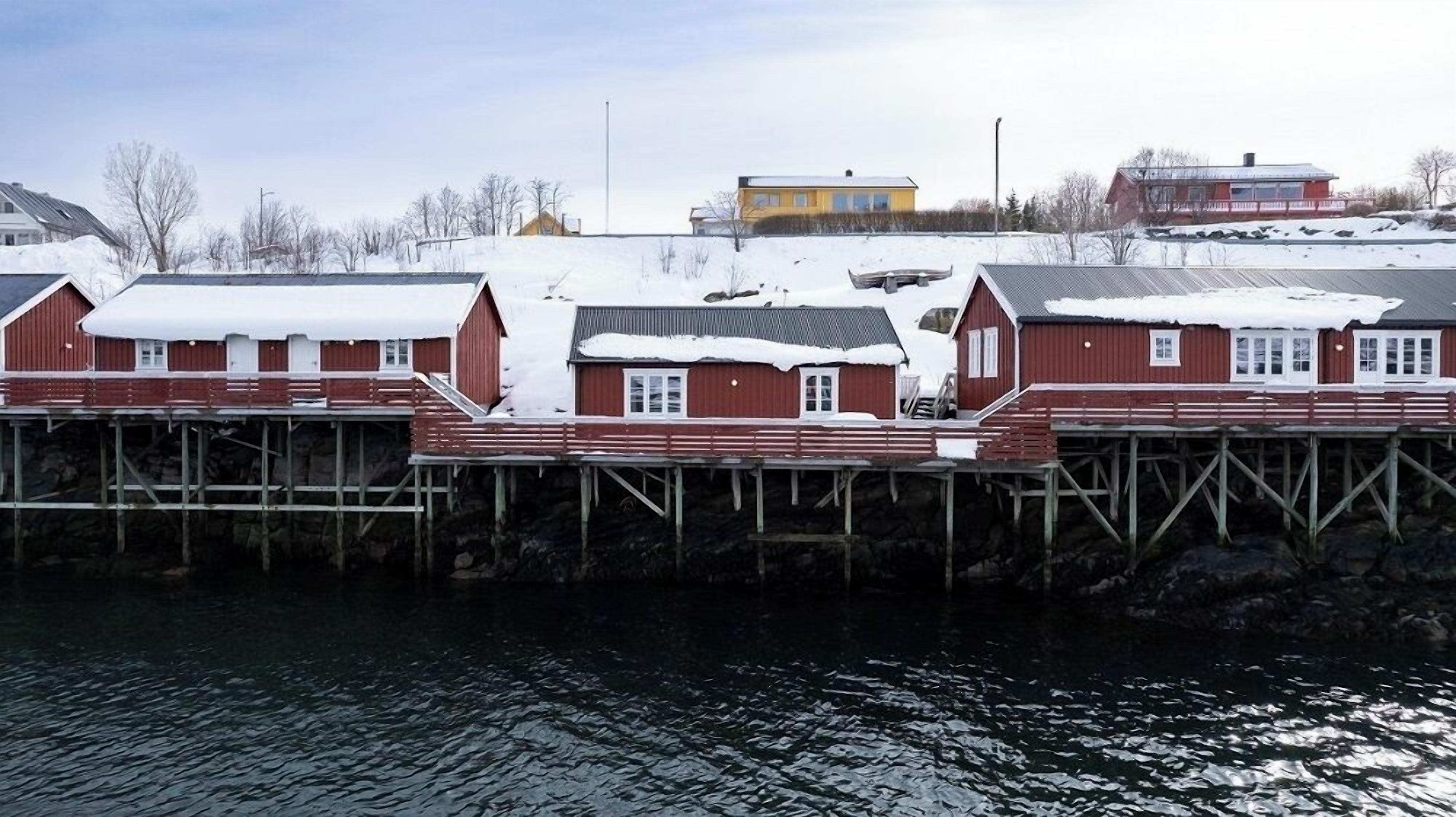 Un edificio frente al agua con fachada roja y detalles blancos, que cuenta con varias ventanas y un muelle que se adentra en el agua.