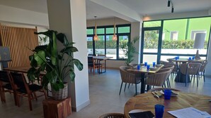 Un restaurante moderno con mesas redondas de madera y sillas de mimbre, grandes ventanales con vistas al verde exterior y plantas en macetas que complementan la decoración.
