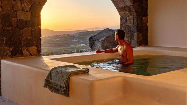 A man in a bathtub with a view of a landscape through a stone archway. - Patina Vivera Estate (Santorini)