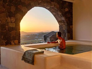 A man in a bathtub with a view of a landscape through a stone archway. - Patina Vivera Estate (Santorini)