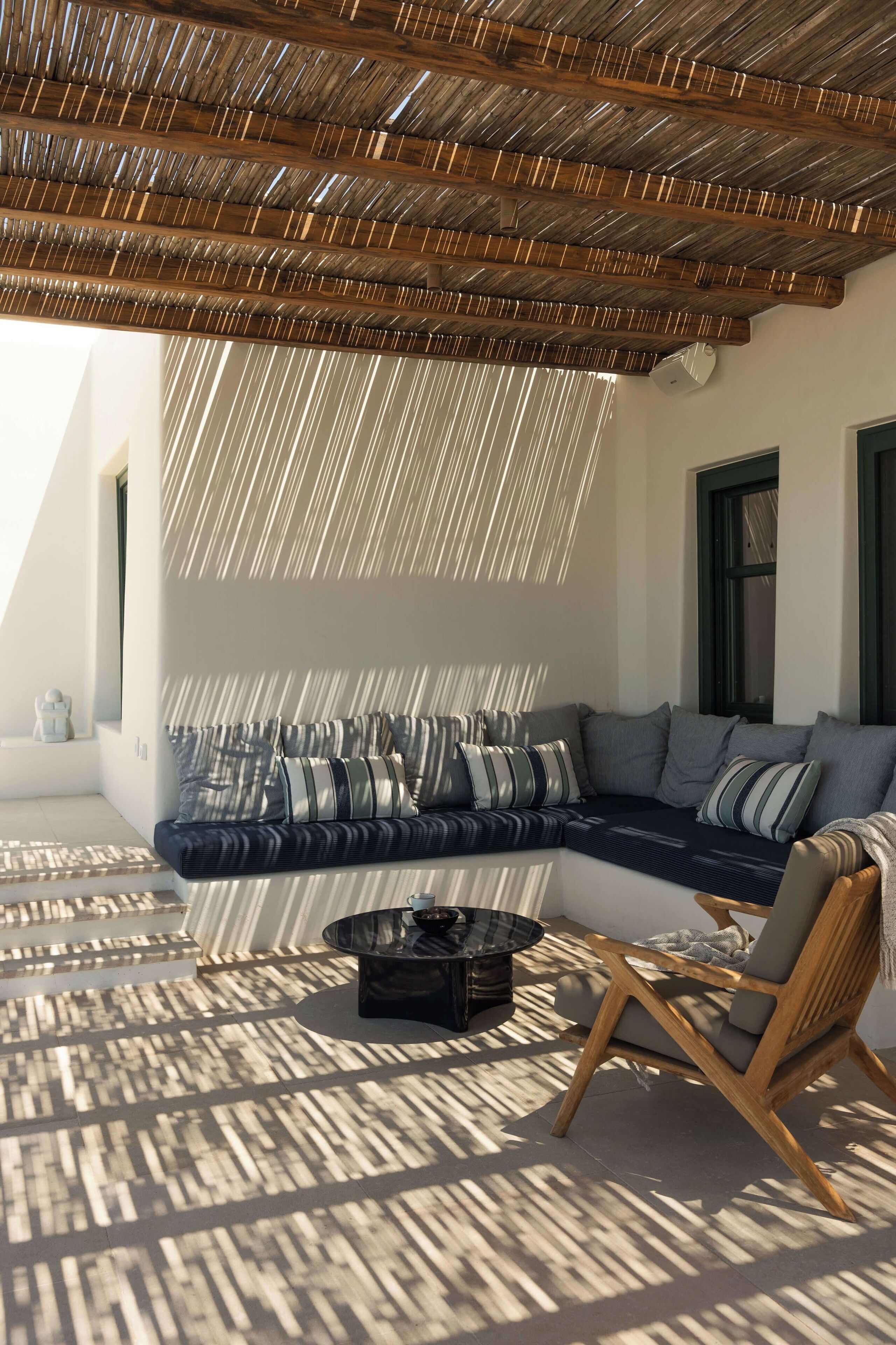 Un espacio al aire libre iluminado por el sol, con techo de madera, un sofá con cojines a rayas y una silla de madera.