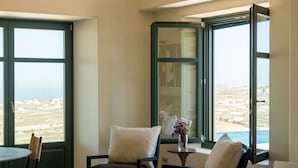 A spacious room with a large window offering a view of the sea, a comfortable seating area with a table, and a small decorative vase. - Patina Vivera Estate (Santorini)