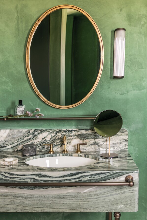 Un baño con lavamanos de mármol, grifería dorada, un espejo ovalado grande y pared verde.