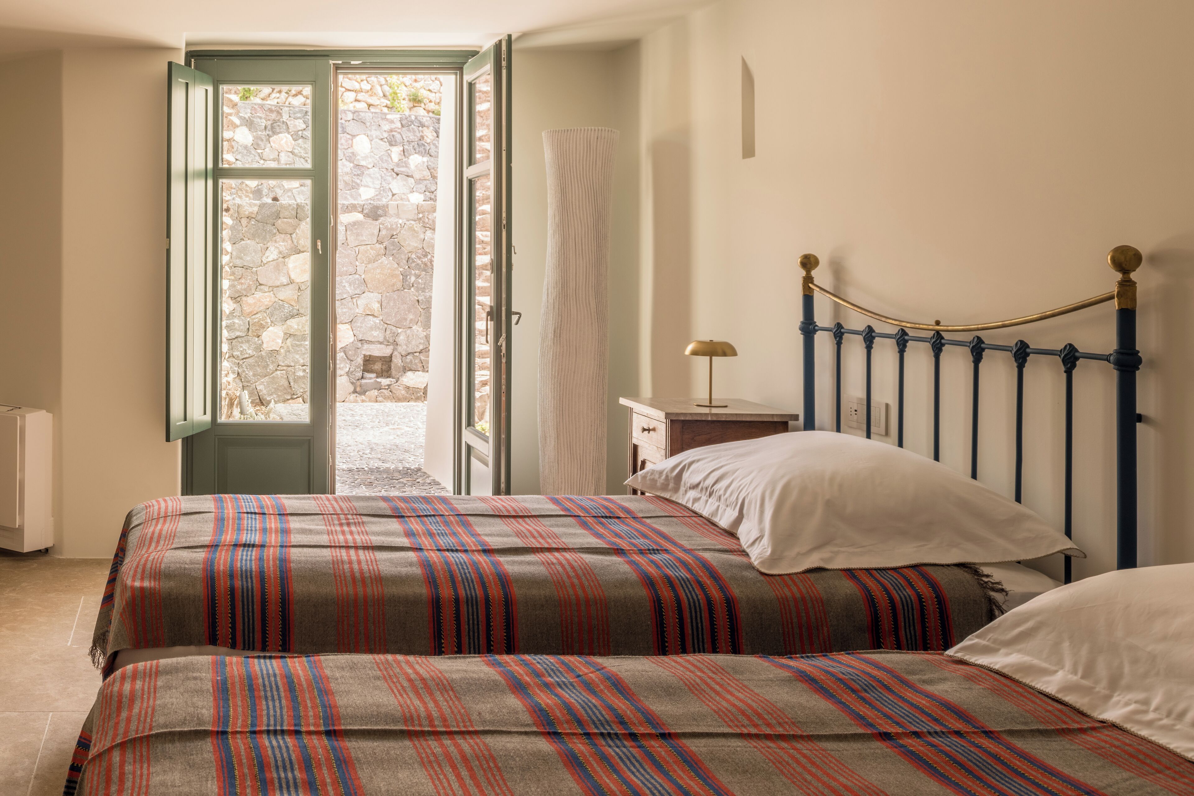 A bedroom with two beds, a stone wall, and a view through an open door.