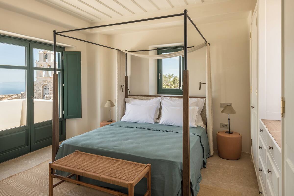 A four-poster bed with a view of a white tower through a window, a bedside table, a lamp, and a wooden bench.