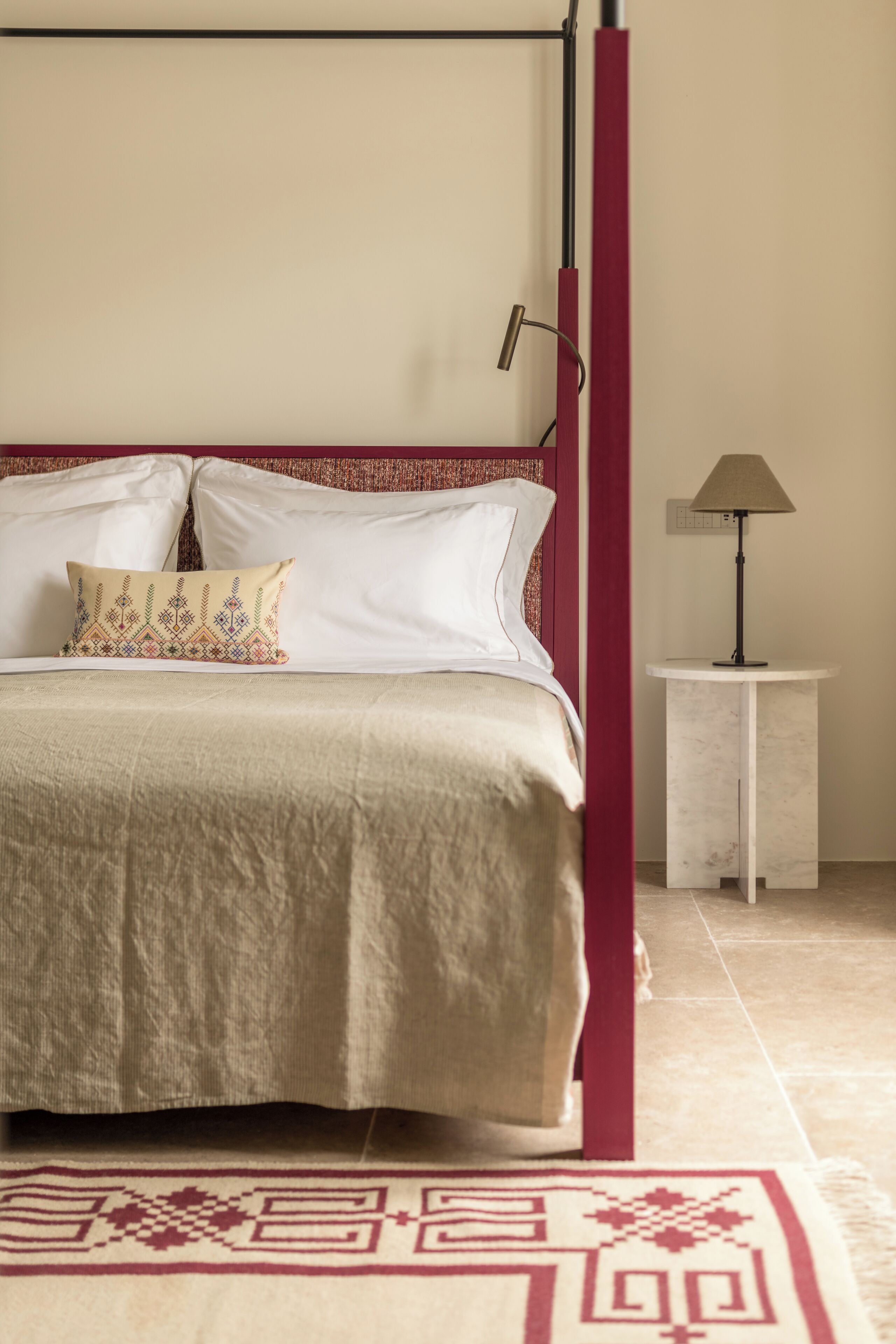 A neatly made bed with a beige bedspread and two pillows, a small bedside table with a lamp, and a patterned rug on the floor.