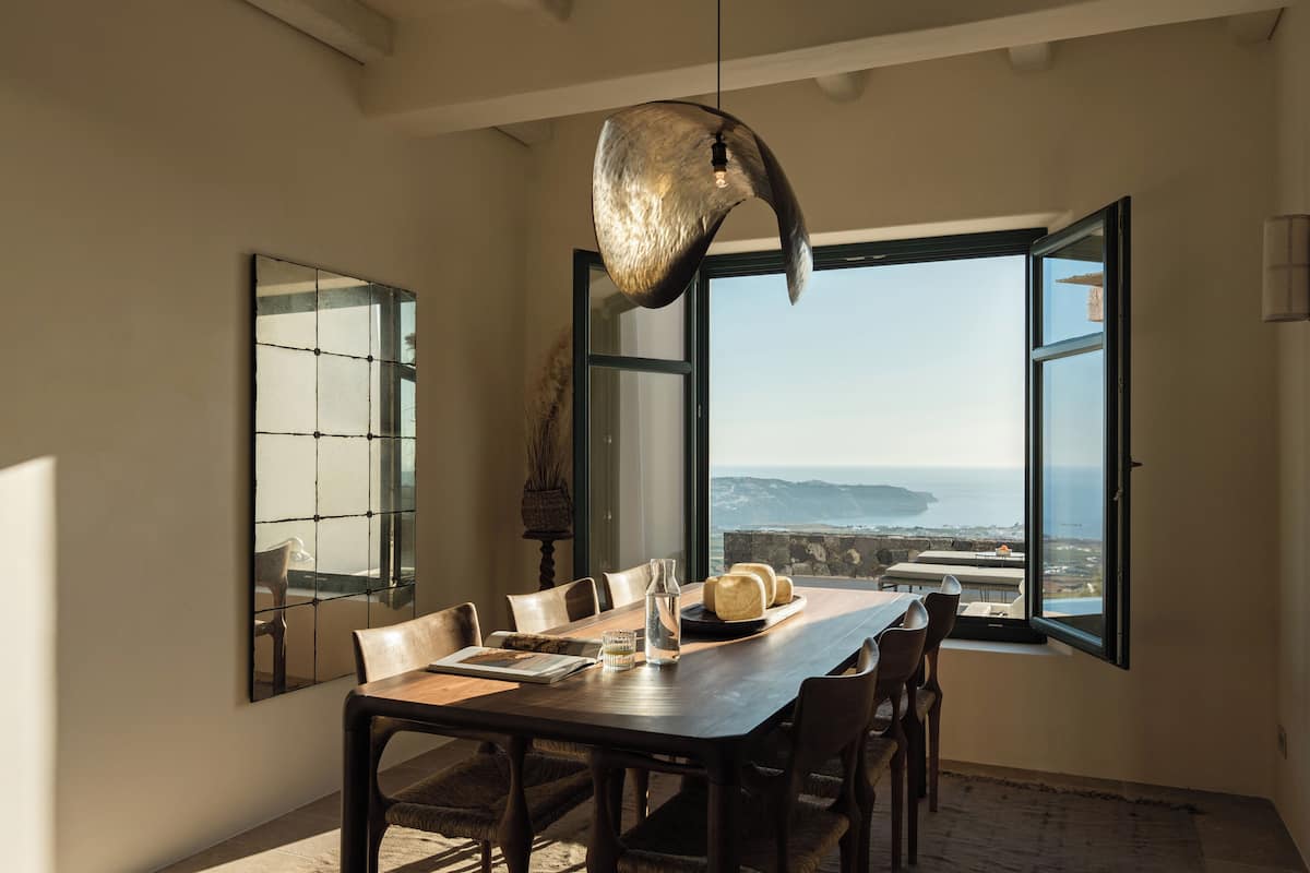 A dining area with a large table, chairs, a hanging light fixture, a window with an open door, and a view of the sea.