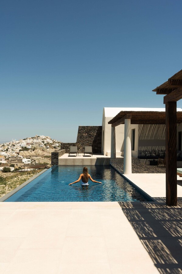 A swimming pool with a person in the water, a white building with a stone facade, and a view of a hill with white buildings. - Patina Vivera Estate (Santorini)