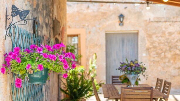 A rustic outdoor dining area with a stone wall, wooden table and chairs, and hanging flower baskets. - Can Fiolet in Algaida (Algaida)