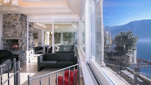 A modern living room with a stone fireplace, a glass door leading to a balcony, and a view of a lake and mountains. - Villa Lugano Lakefront (Valsolda)
