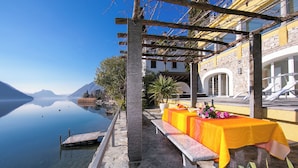 A lakeside terrace with a dining table set for two, overlooking a calm lake and mountains. - Villa Lugano Lakefront (Valsolda)