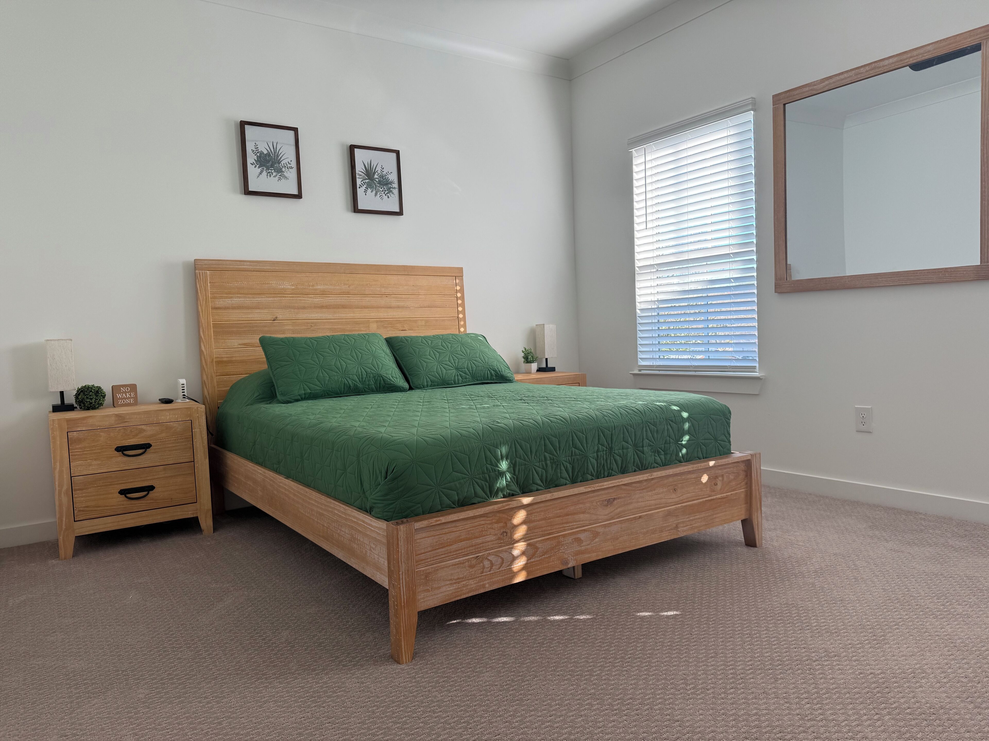 Una camera da letto con un letto in legno, biancheria verde, un comodino con cassetti, una lampada da comodino e una finestra con persiane.
