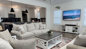 A modern living room with a grey sofa, white armchairs, a coffee table, and a TV on the wall. - Large Family Friendly 4BR/10 Bed. Pool, Bay Access (Orange Beach)
