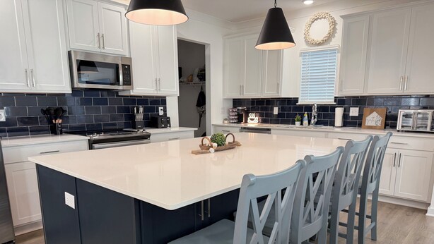 A modern kitchen with white cabinets, a large island with bar stools, and black and white tile backsplash. - Large Family Friendly 4BR/10 Bed. Pool, Bay Access (Orange Beach)
