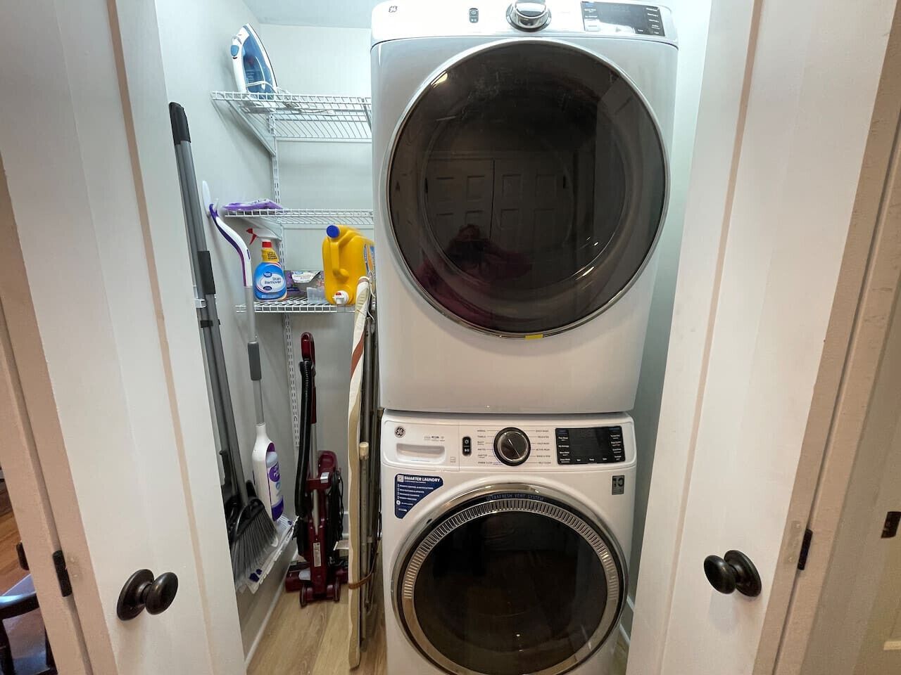 A compact laundry room with a stacked washer and dryer, shelves with cleaning supplies, and a broom.
