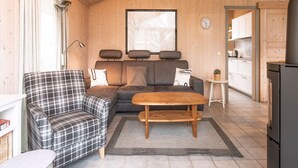 L’intérieur d’un chalet accueillant, avec un canapé, un fauteuil à motifs, une table basse en bois, un coin cuisine et une grande fenêtre agrémentée de rideaux.