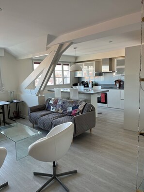 Un espacio moderno para vivir con sofá, una mesa de centro de vidrio, una silla blanca y una zona de cocina con armarios y una mesa de comedor.