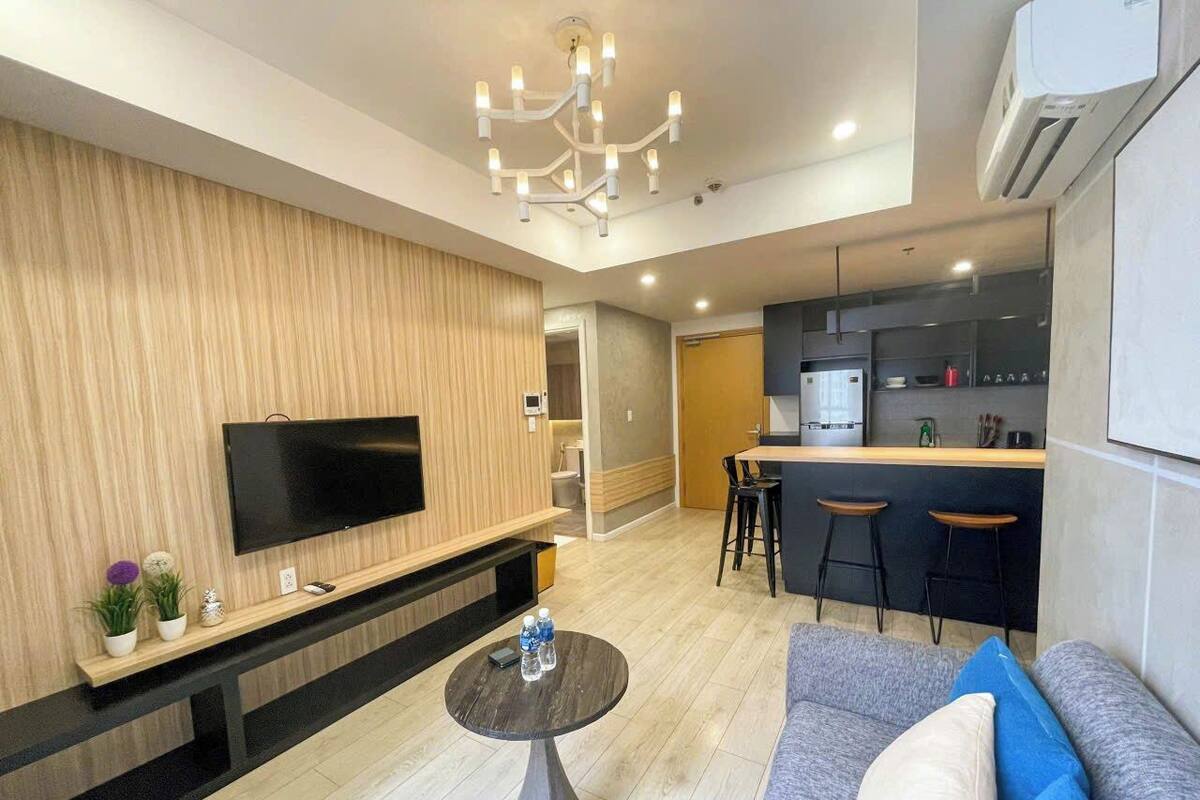 Uma sala de estar moderna com uma TV de tela plana, uma mesa de centro redonda e um sofá com almofadas. Uma área de cozinha com balcão e banquetas é visível ao fundo.