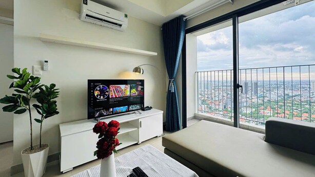 Ein modernes Wohnzimmer mit Flachbildfernseher, einer weißen Unterhaltungsecke, einer Topfpflanze, einer Vase mit roten Blumen, einer Fernbedienung und einem großen Fenster mit Blick auf die Stadt.