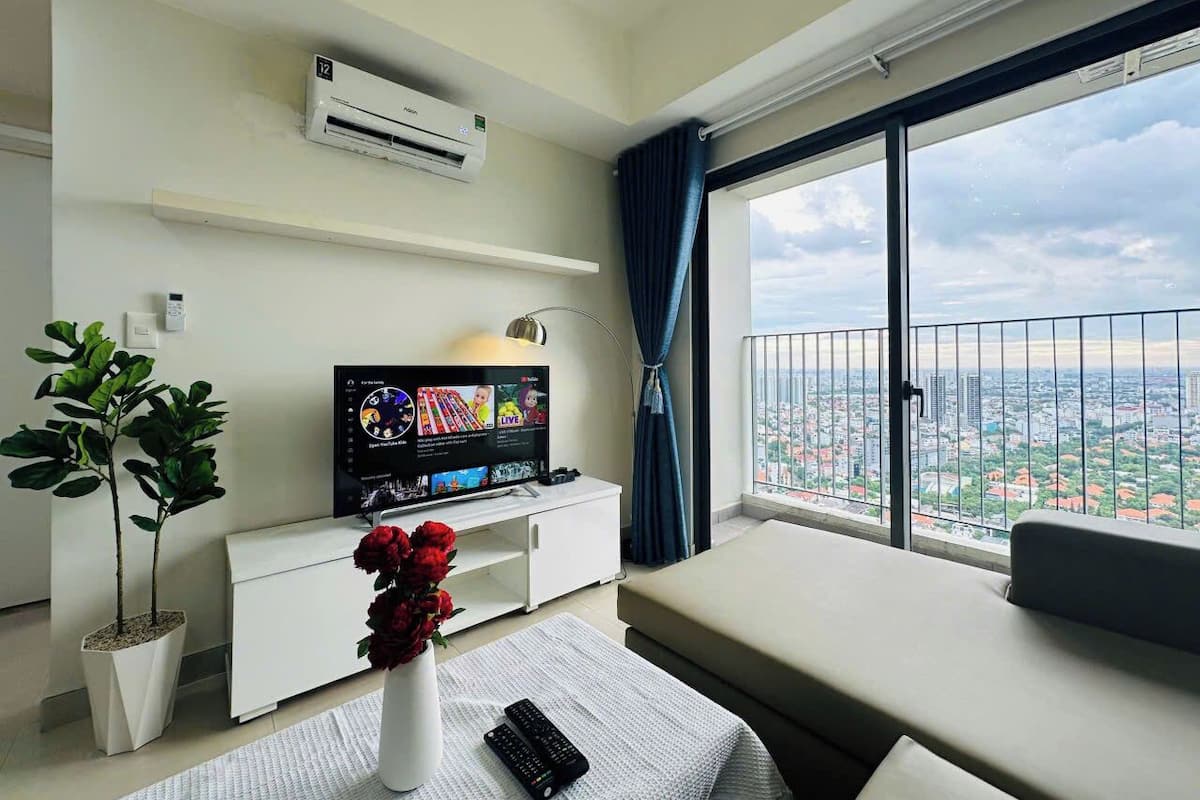Uma sala de estar moderna com uma TV de tela plana, um móvel branco para eletrônicos, uma planta em vaso, um vaso com flores vermelhas, um controle remoto e uma grande janela com vista para a cidade.