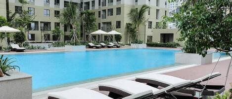 A swimming pool with lounge chairs and umbrellas, surrounded by palm trees and a high-rise building.