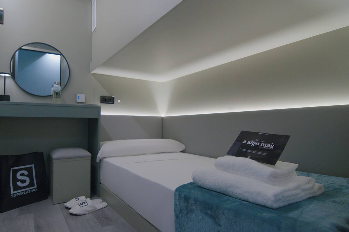 A modern, compact hotel room with a single bed, a small bedside table, a mirror, and a wall-mounted light fixture.