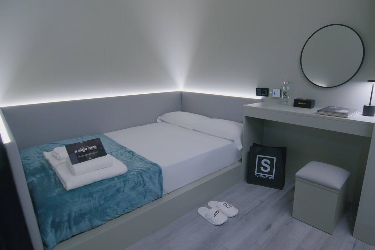 A compact hotel room with a single bed, a bedside table with a lamp, a small mirror, and a bag of toiletries.