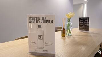 A hotel room with a wooden table, a poster advertising a water bottle, a vase with flowers, and a menu board.