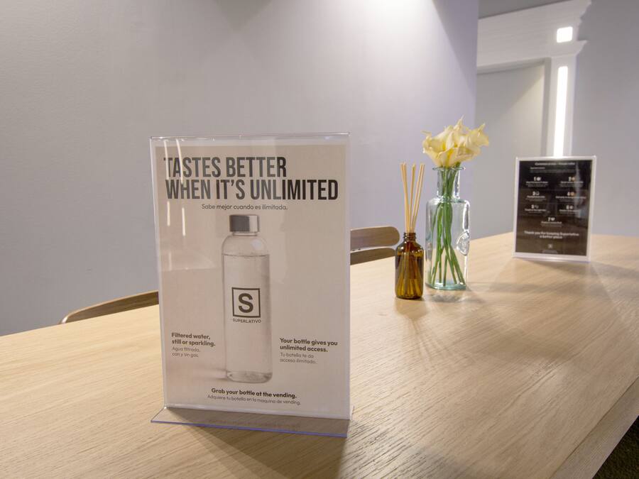 A hotel room with a wooden table, a poster advertising a water bottle, a vase with flowers, and a menu board.