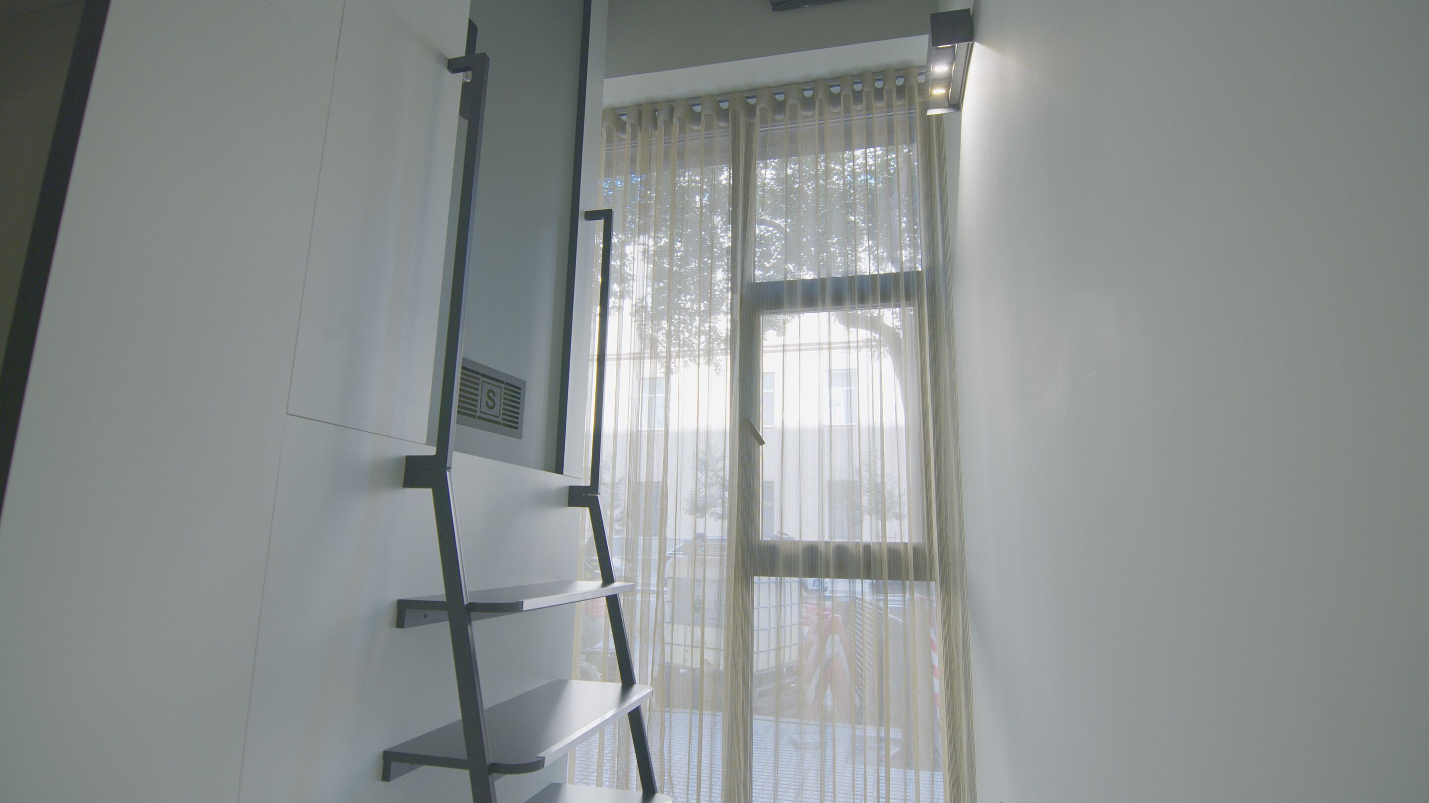 Una habitación con una ventana cubierta por cortinas translúcidas, una escalera metálica y una rejilla de ventilación en la pared.