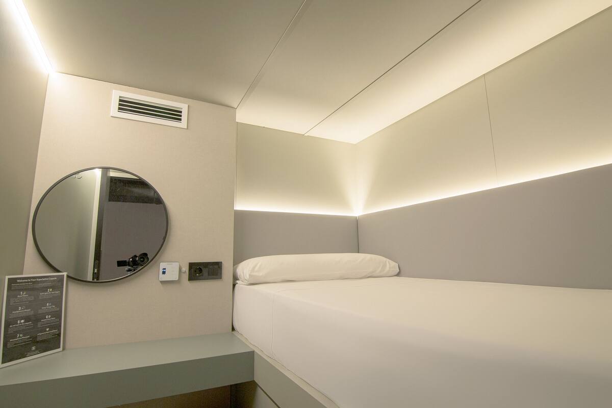 A modern hotel room with a bed, a round mirror, a wall-mounted air vent, and a small table with a sign.